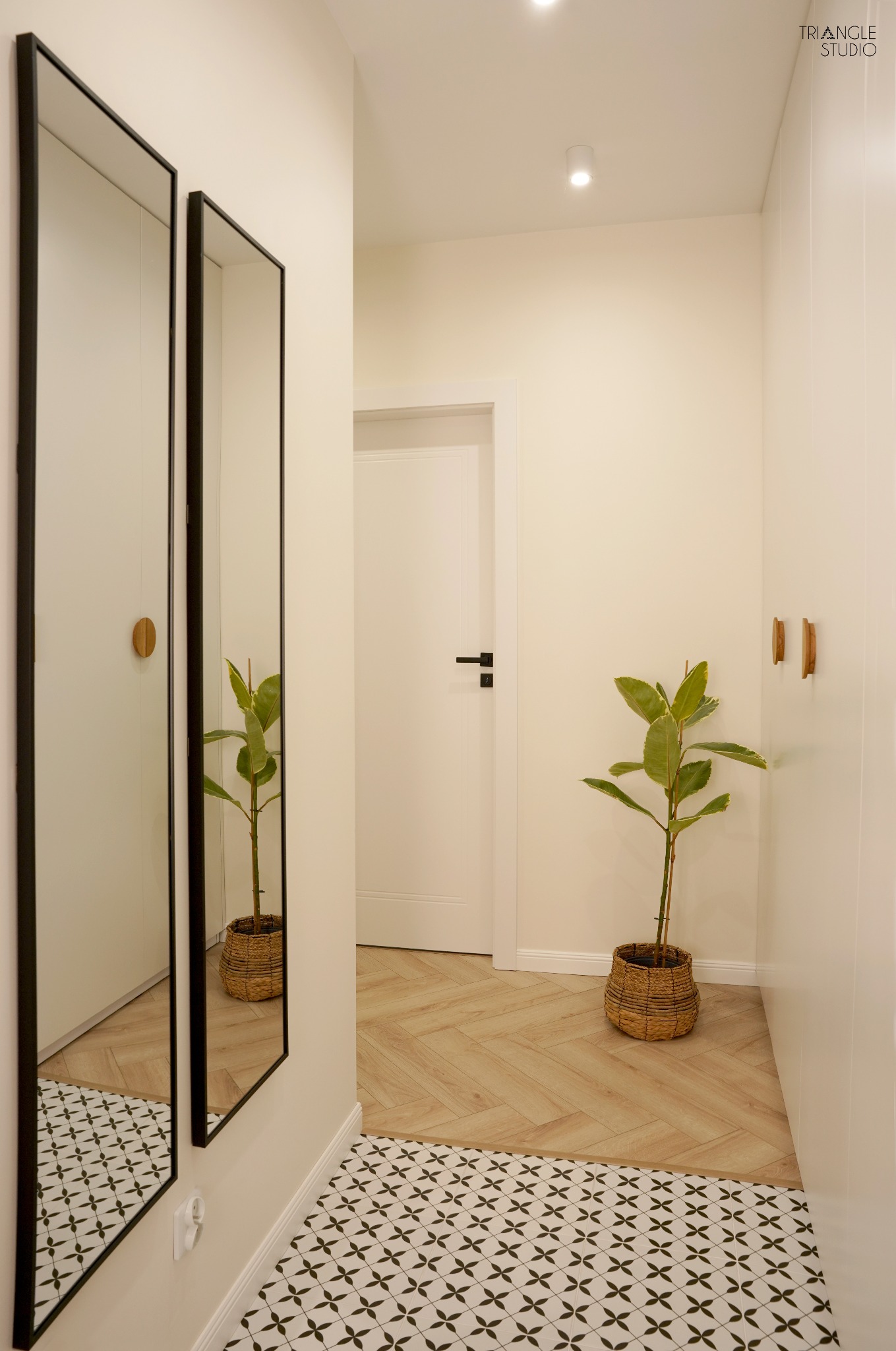 Jasny korytarz z dwoma lustrami w czarnych ramach, rośliną w wiklinowym koszu i wzorzystą podłogą. Widoczne drewniane uchwyty i białe drzwi.