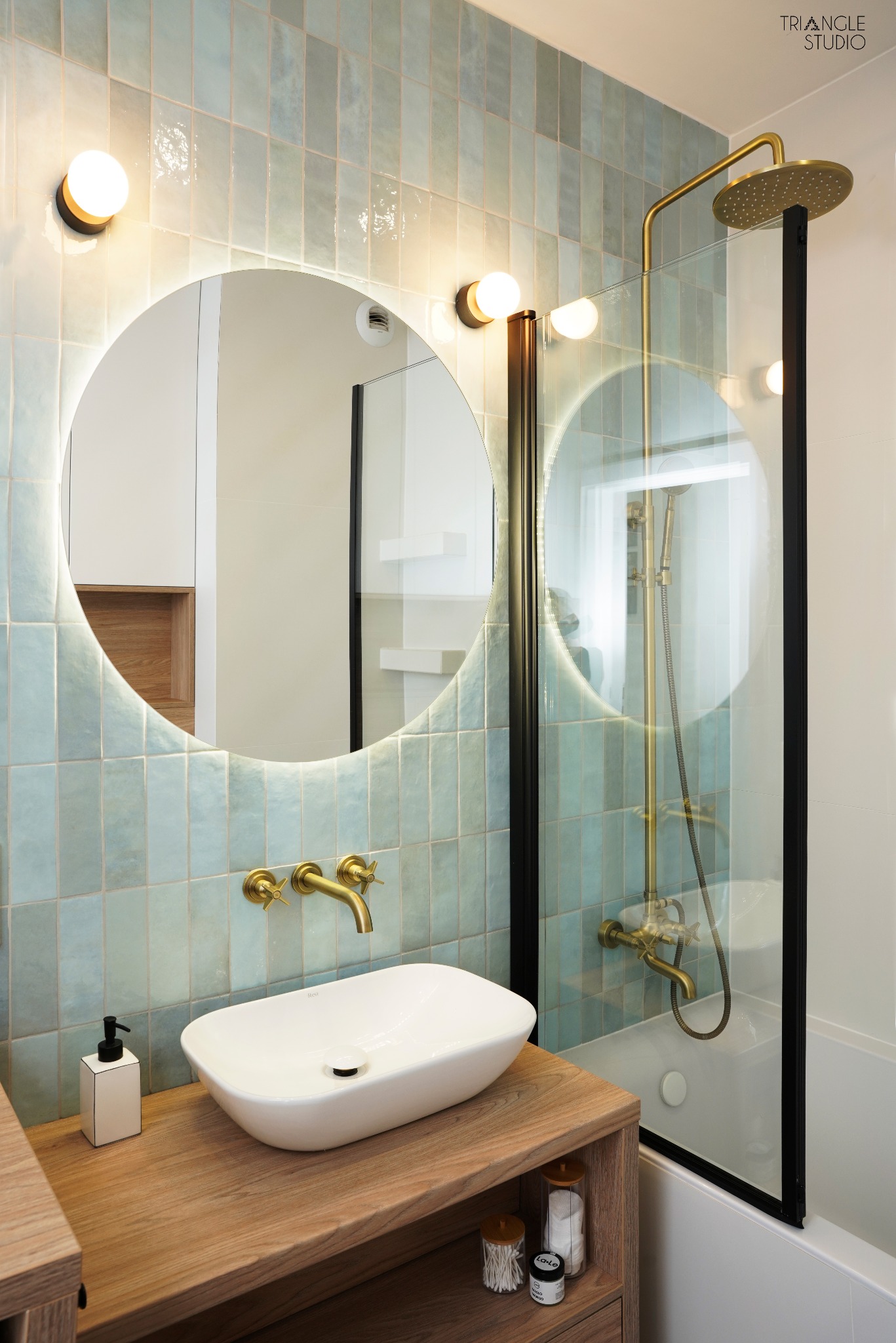 Nowoczesna łazienka z owalną umywalką, złotą armaturą i podświetlanym lustrem. Błękitne, pionowe płytki na ścianie. Kabina prysznicowa ze złotym zestawem natryskowym.