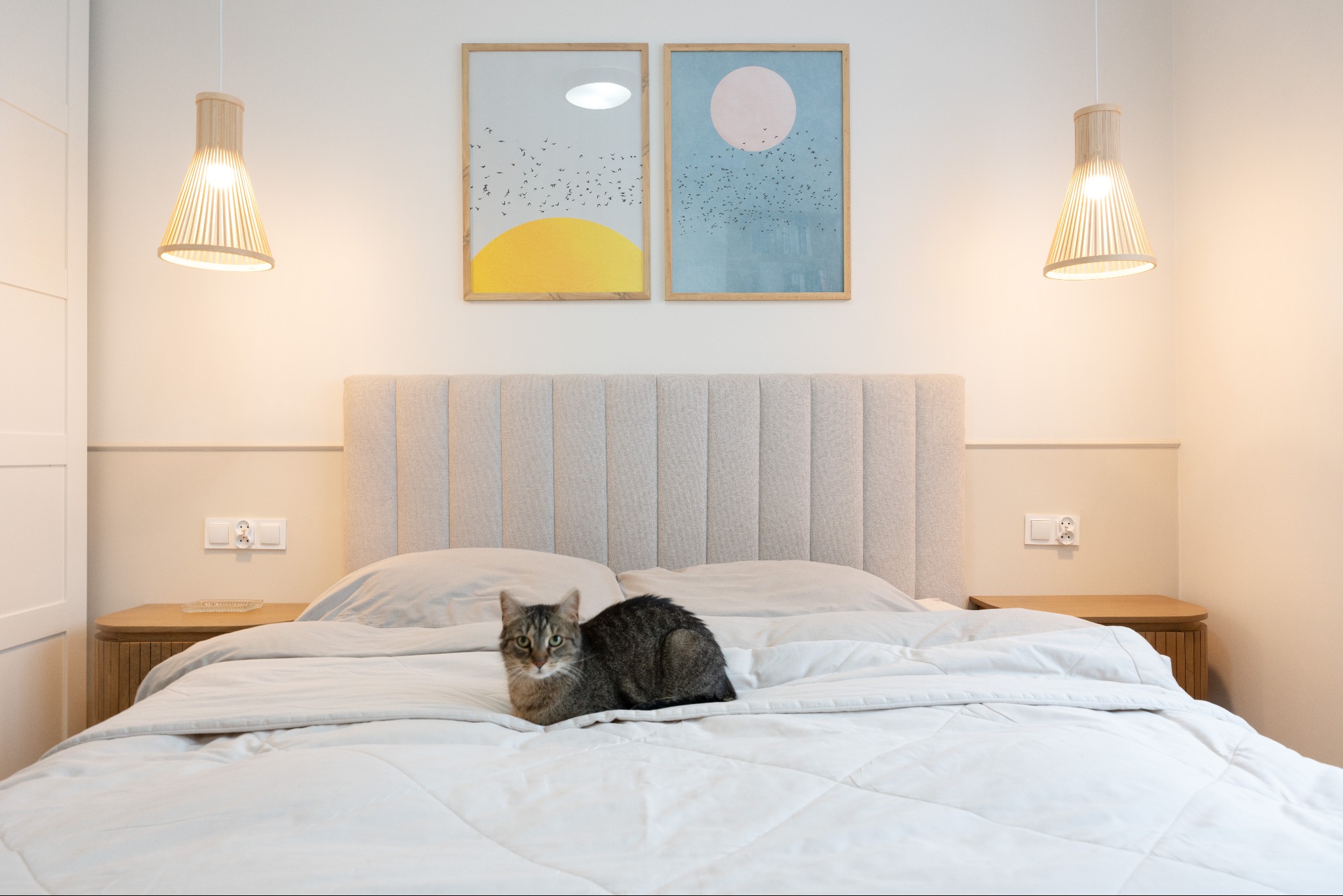Jasna sypialnia w neutralnych barwach z łóżkiem, na którym leży pręgowany kot. Nad łóżkiem wiszą dwa abstrakcyjne obrazy i lampy z drewnianymi abażurami.