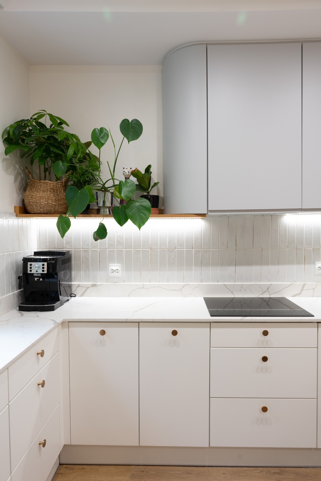 Jasna kuchnia z białymi szafkami, blatem imitującym marmur i roślinami doniczkowymi na półce. Ekspres do kawy na blacie. Minimalistyczny design z akcentami drewna.