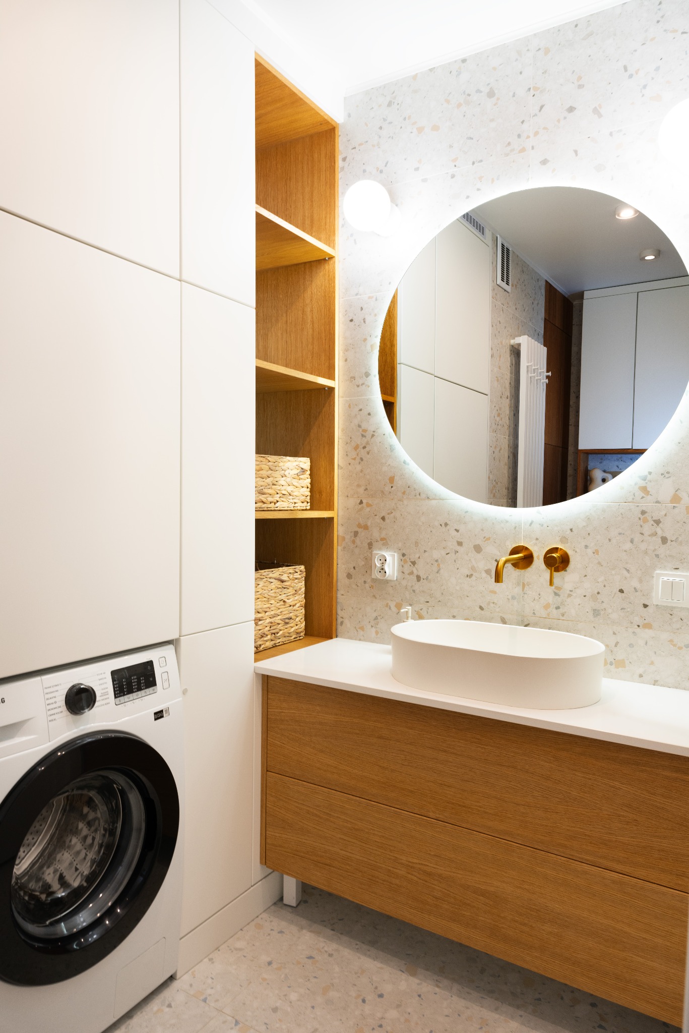 Nowoczesna łazienka z okrągłym lustrem podświetlanym LED, umywalką nablatową, złotą armaturą i pralką. Jasne drewno szafek i terazzo na ścianie.