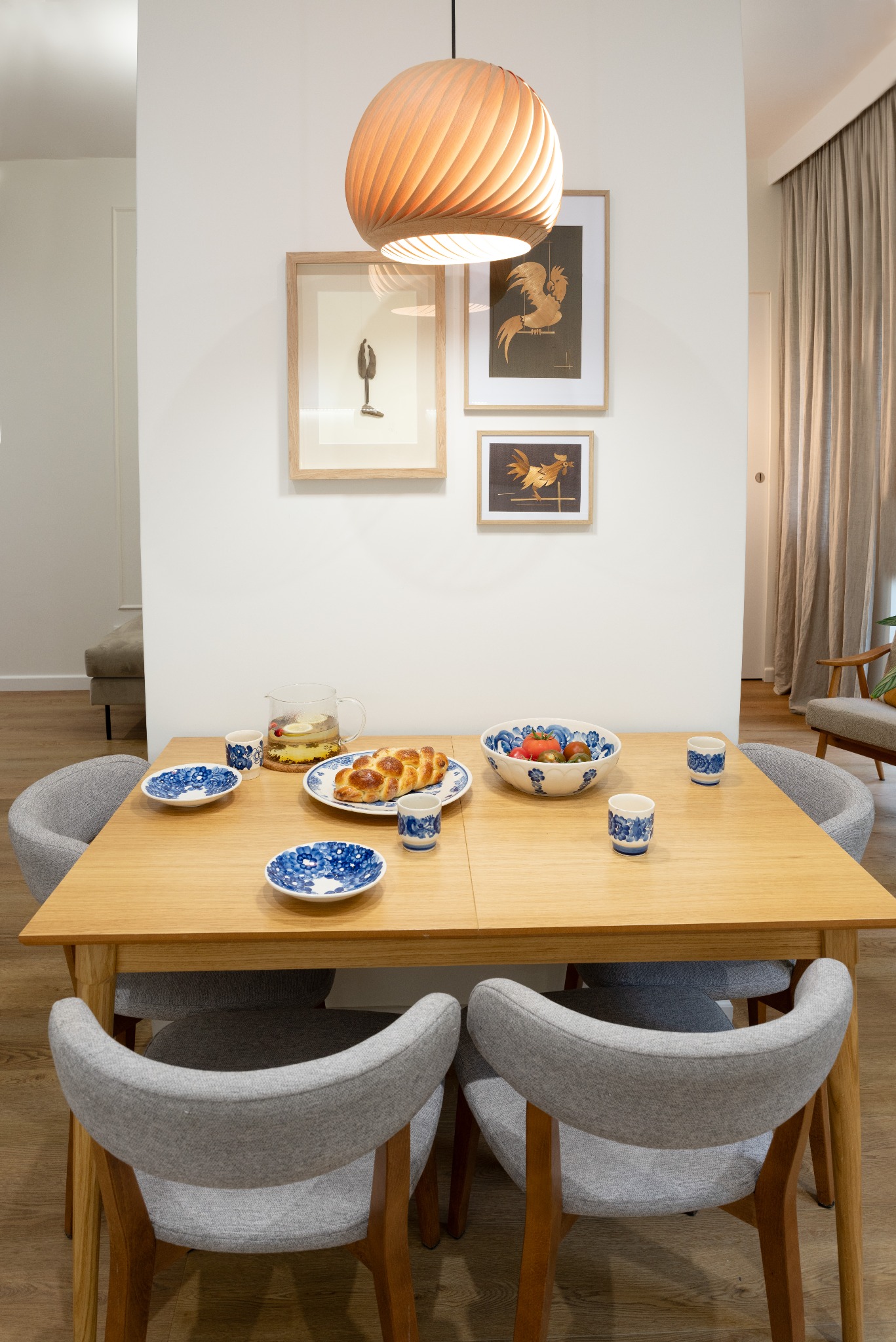 Jasne drewno stołu jadalnego z szarymi krzesłami, nakryte niebieską porcelaną. W tle grafiki na ścianie i lampa z drewnianych listewek. Naturalne światło.