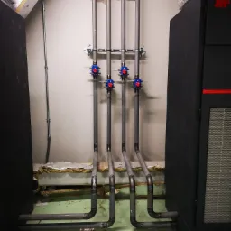 Cztery pionowe rury instalacyjne z niebiesko-czerwonymi zaworami zamocowane do ściany, przechodzące przez otwór w suficie, podłączone do czarnej jednostki po prawej stronie, widoczne ślady spawania...