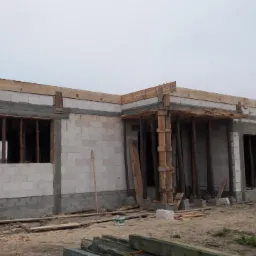 Surowy stan parterowego budynku z betonu komórkowego w trakcie budowy, widoczne drewniane szalunki okienne i stropowe, teren budowy z materiałami.