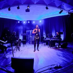 Zespół muzyczny na scenie podczas występu, wokalistka w czarnej sukience, oświetlenie sceniczne w odcieniach niebieskiego, widoczni muzycy grający na instrumentach klawiszowych i perkusji.