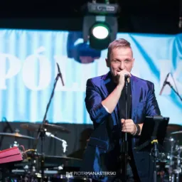 Wokalista w błyszczącym niebieskim garniturze śpiewa do mikrofonu na scenie z perkusją i klawiszami w tle.