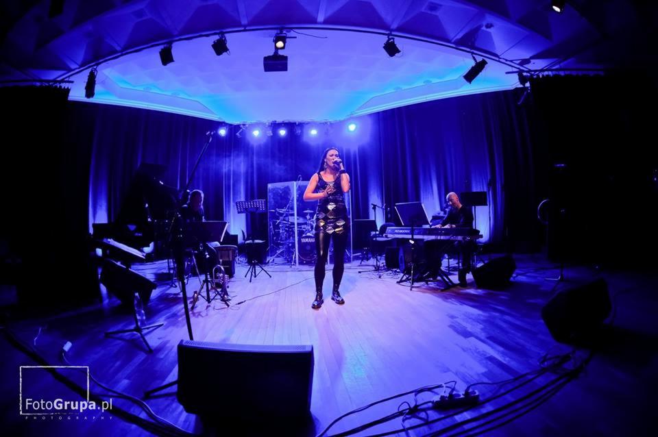 Zespół muzyczny na scenie podczas występu, wokalistka w czarnej sukience, oświetlenie sceniczne w odcieniach niebieskiego, widoczni muzycy grający na instrumentach klawiszowych i perkusji.