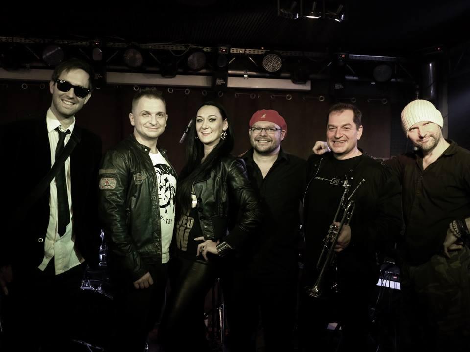 Sześcioosobowy zespół na scenie w ciemnym oświetleniu, z trąbką w ręku jednego z muzyków, w różnorodnych strojach, w tym skórzana kurtka i garnitur z krawatem.