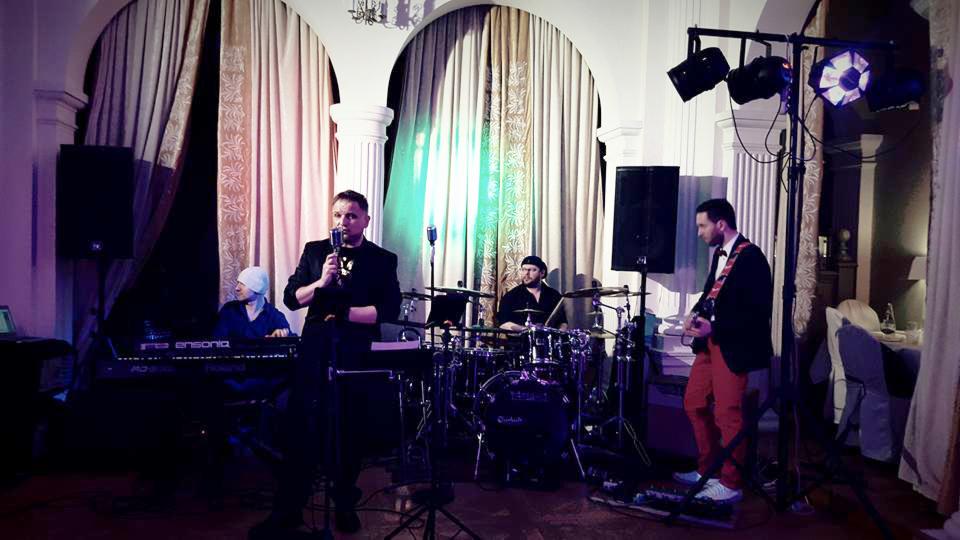 Występujący na scenie zespół muzyczny: wokalista z mikrofonem, klawiszowiec w białej czapce, perkusista i gitarzysta w czerwonych spodniach, oświetlenie sceniczne