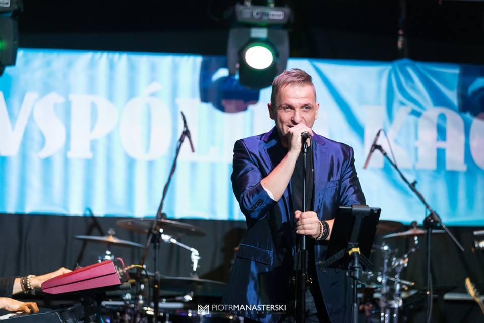 Wokalista w błyszczącym niebieskim garniturze śpiewa do mikrofonu na scenie z perkusją i klawiszami w tle.