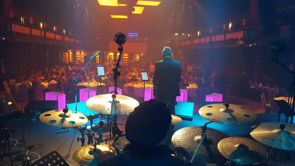 Perkusista z widokiem na scenę i publiczność podczas występu na eleganckim przyjęciu z oświetleniem w kolorze fioletowym i pomarańczowym.