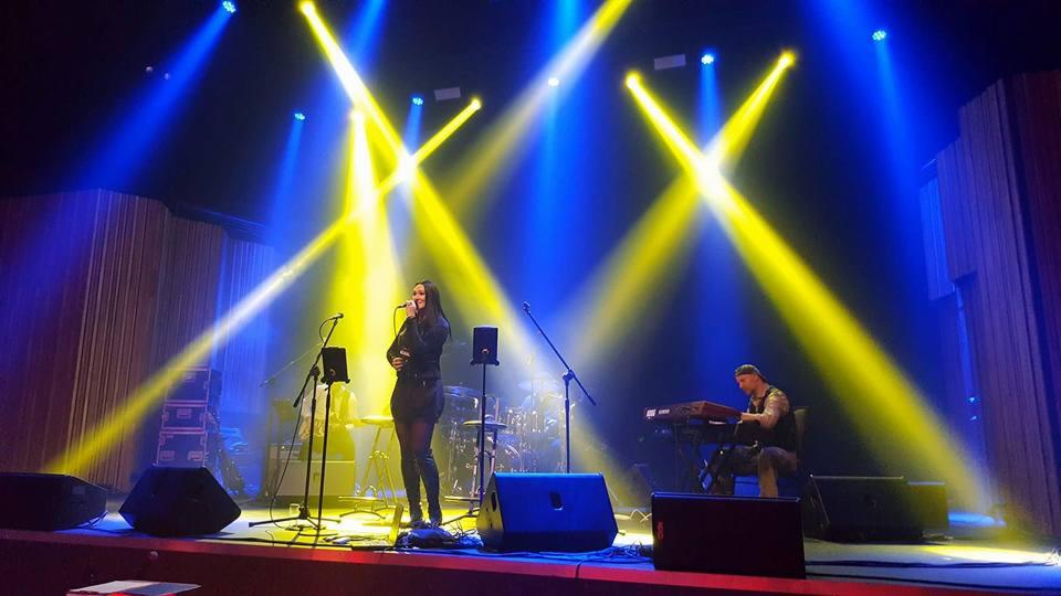Wokalistka w czarnej sukience śpiewa do mikrofonu na scenie, obok klawiszowiec w kamuflażu, scena oświetlona niebieskimi i żółtymi reflektorami.
