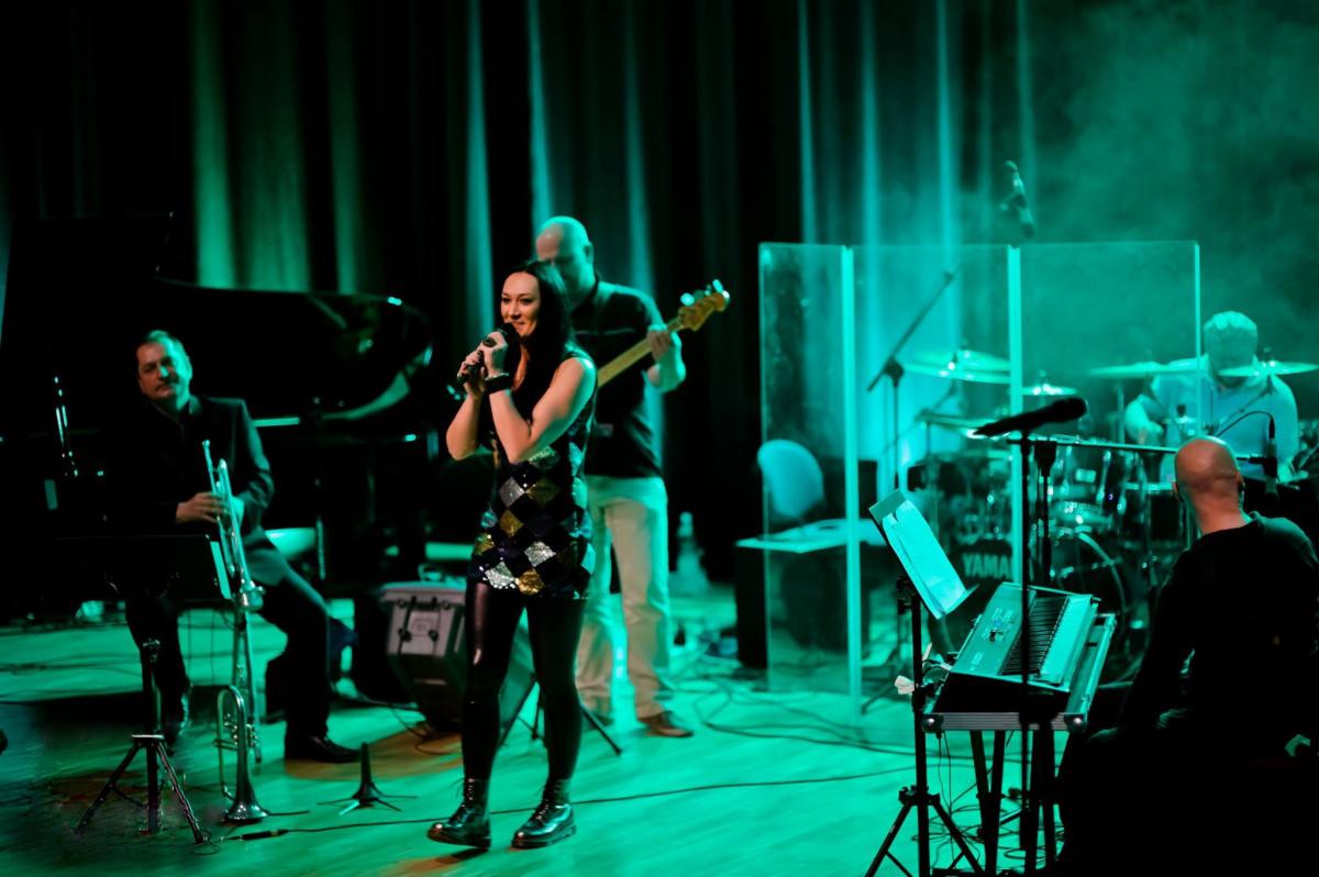 Wokalistka w cekinowej sukience i czarnych legginsach śpiewa do mikrofonu na scenie oświetlonej zielonym światłem, z zespołem grającym na instrumentach w tle.