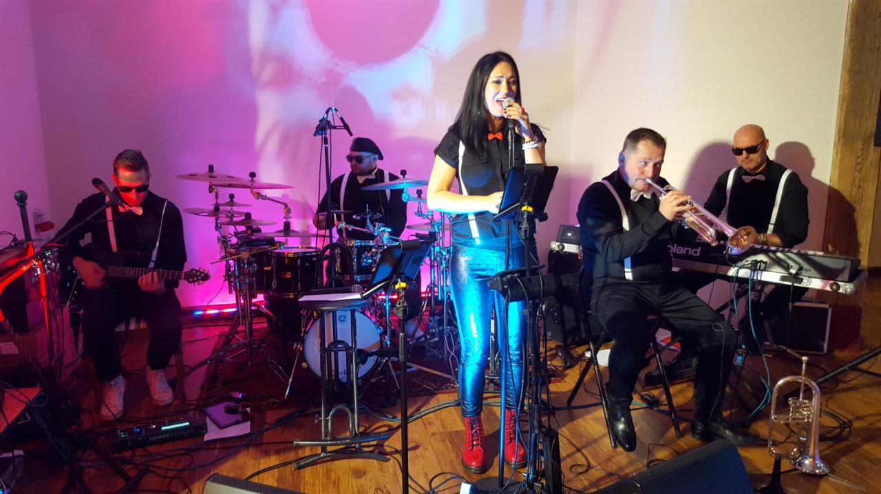 Zespół muzyczny w trakcie występu: wokalistka w błyszczących spodniach i czerwonych butach śpiewa do mikrofonu, gitarzysta gra na gitarze, perkusista za bębnami, a dwóch muzyków gra na trąbce...