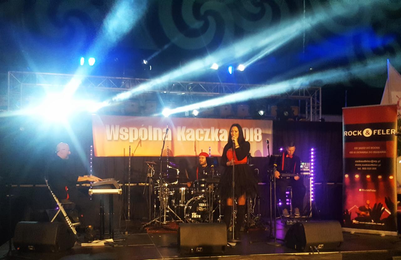 Zespół muzyczny na scenie oświetlonej reflektorami, wokalistka śpiewa do mikrofonu, w tle baner z napisem 'Wspólna Kaczka 2018', po prawej stronie baner reklamowy 'Rock&Feler'.