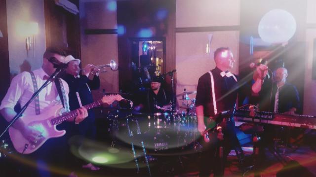 Zespół muzyczny w trakcie występu na scenie: gitarzysta w białej koszuli i szelkach, trębacz w kapeluszu, perkusista za zestawem perkusyjnym Yamaha, klawiszowiec przy keyboardzie Roland.