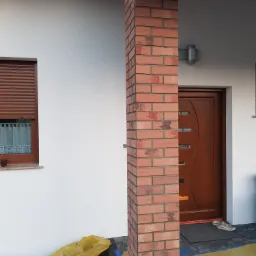 Murowany z cegły filar przy wejściu do domu, zabezpieczony folią malarską, widoczna stolarka okienna z roletami zewnętrznymi i brązowe drzwi wejściowe.
