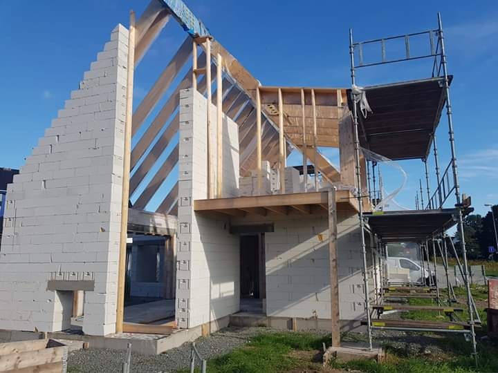 Budowa domu jednorodzinnego w stanie surowym otwartym: ściany z białego bloczka, drewniana konstrukcja dachu, rusztowanie przy ścianie bocznej, błękitne niebo.