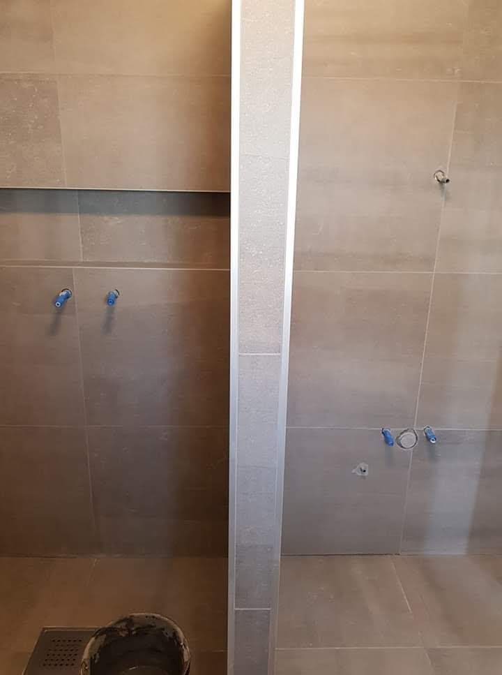 Dwa prysznice w trakcie wykańczania, widoczne szare płytki na ścianach, odpływ liniowy i wiadro z zaprawą na podłodze, przyłącza wodne zabezpieczone niebieskimi zatyczkami.