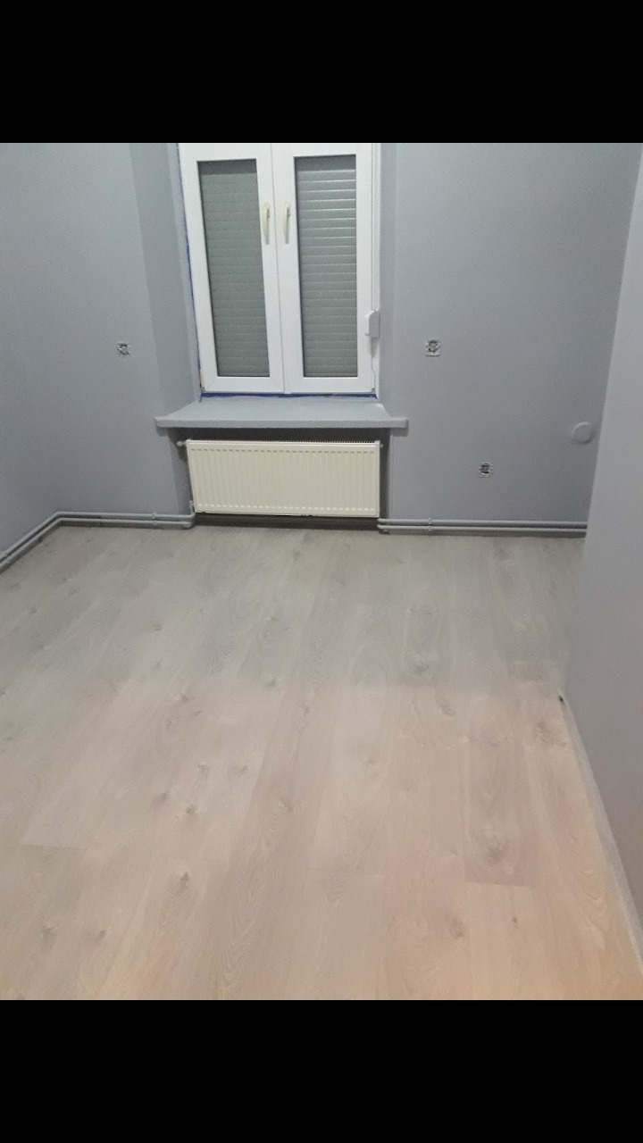 Jasna podłoga panelowa, szare ściany z widocznymi otworami po gniazdkach, biały grzejnik pod oknem z żaluzjami.