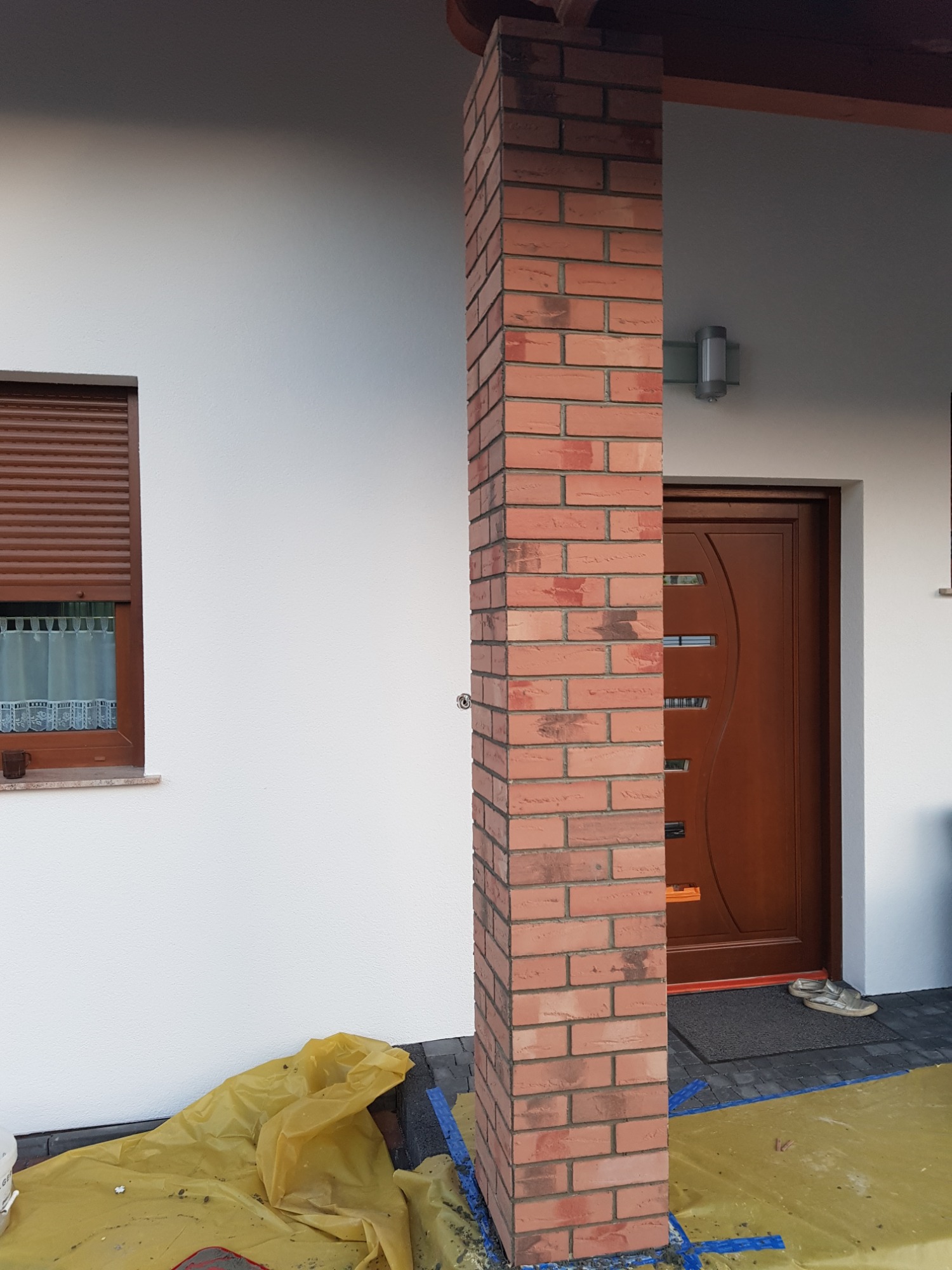 Murowany z cegły filar przy wejściu do domu, zabezpieczony folią malarską, widoczna stolarka okienna z roletami zewnętrznymi i brązowe drzwi wejściowe.