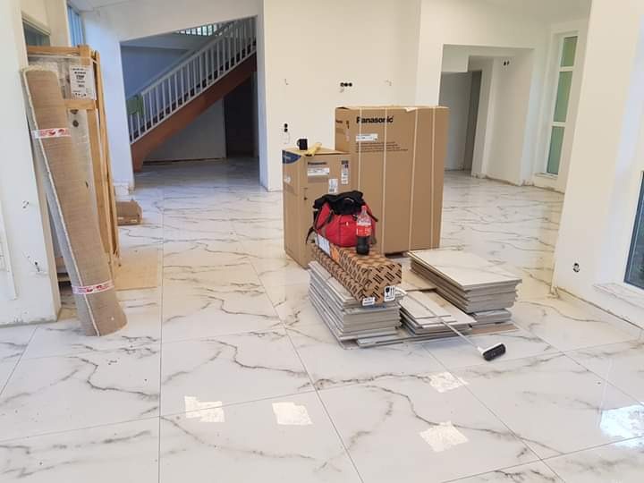 Białe płytki podłogowe z szarym marmurkowym wzorem, ułożone w przestronnym wnętrzu w trakcie remontu, z zapasem płytek, kartonami i zwiniętym dywanem w tle schodów.