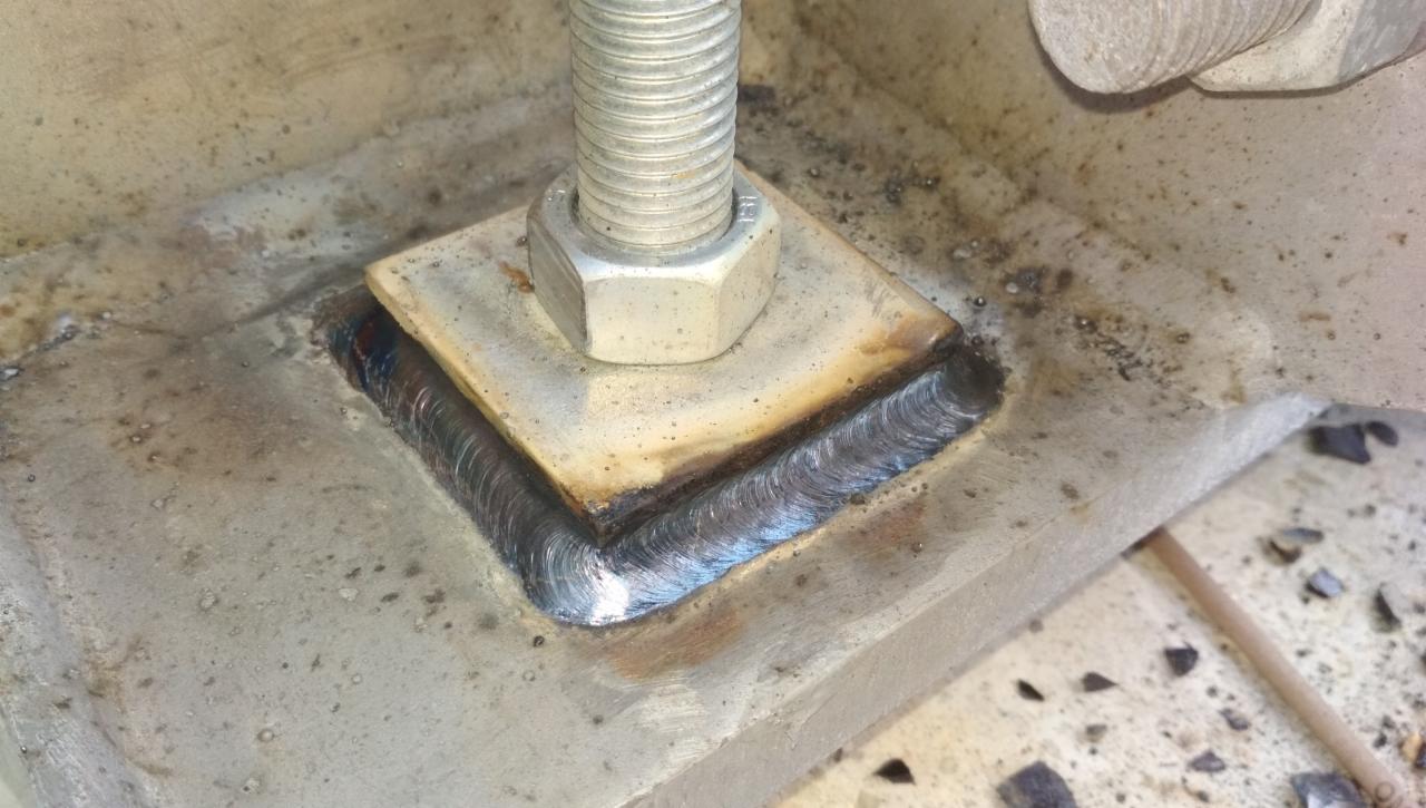 Szczegółowe ujęcie spawu łączącego kwadratową płytkę z grubą metalową konstrukcją, z widoczną śrubą z nakrętką na płytce, ukazujące precyzyjne wykonanie spoiny.