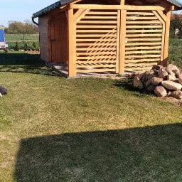 Drewniana konstrukcja z poziomymi listwami, prawdopodobnie wiata lub składzik, z czarnym dachem, stojąca na trawniku, z ułożonymi obok kamieniami.