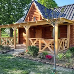 Zewnętrzny widok małego, parterowego domu z bali z zadaszonym tarasem, czarną dachówką i ogrodem z obrzeżami z małych pniaków.