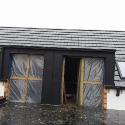 Nowe, grafitowe dachówki ceramiczne na skośnym dachu z oknem dachowym; widoczne zabezpieczone folią okna lukarny i fragment obróbki blacharskiej w kolorze czarnym.