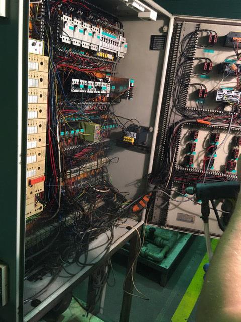 Otwarta szafa sterownicza z widocznymi licznymi przewodami, przekaźnikami i modułami sterującymi, ukazująca skomplikowaną instalację elektryczną w zakładzie przemysłowym.