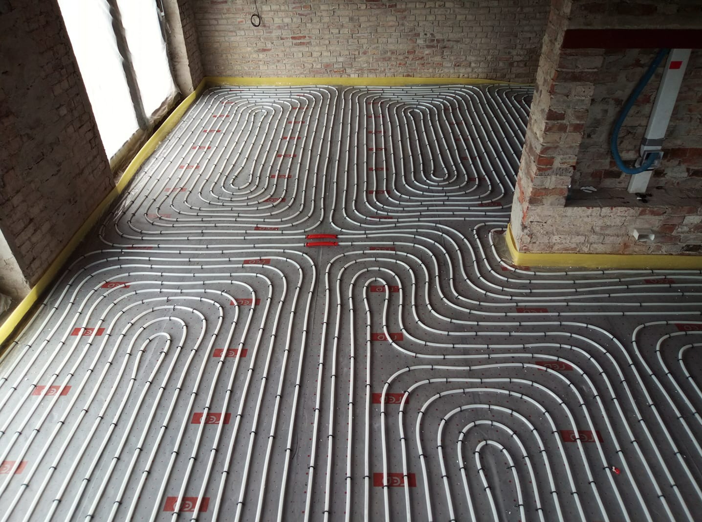 Rozłożone rury ogrzewania podłogowego na szarej macie izolacyjnej w surowym wnętrzu z ceglanymi ścianami, widok z góry.