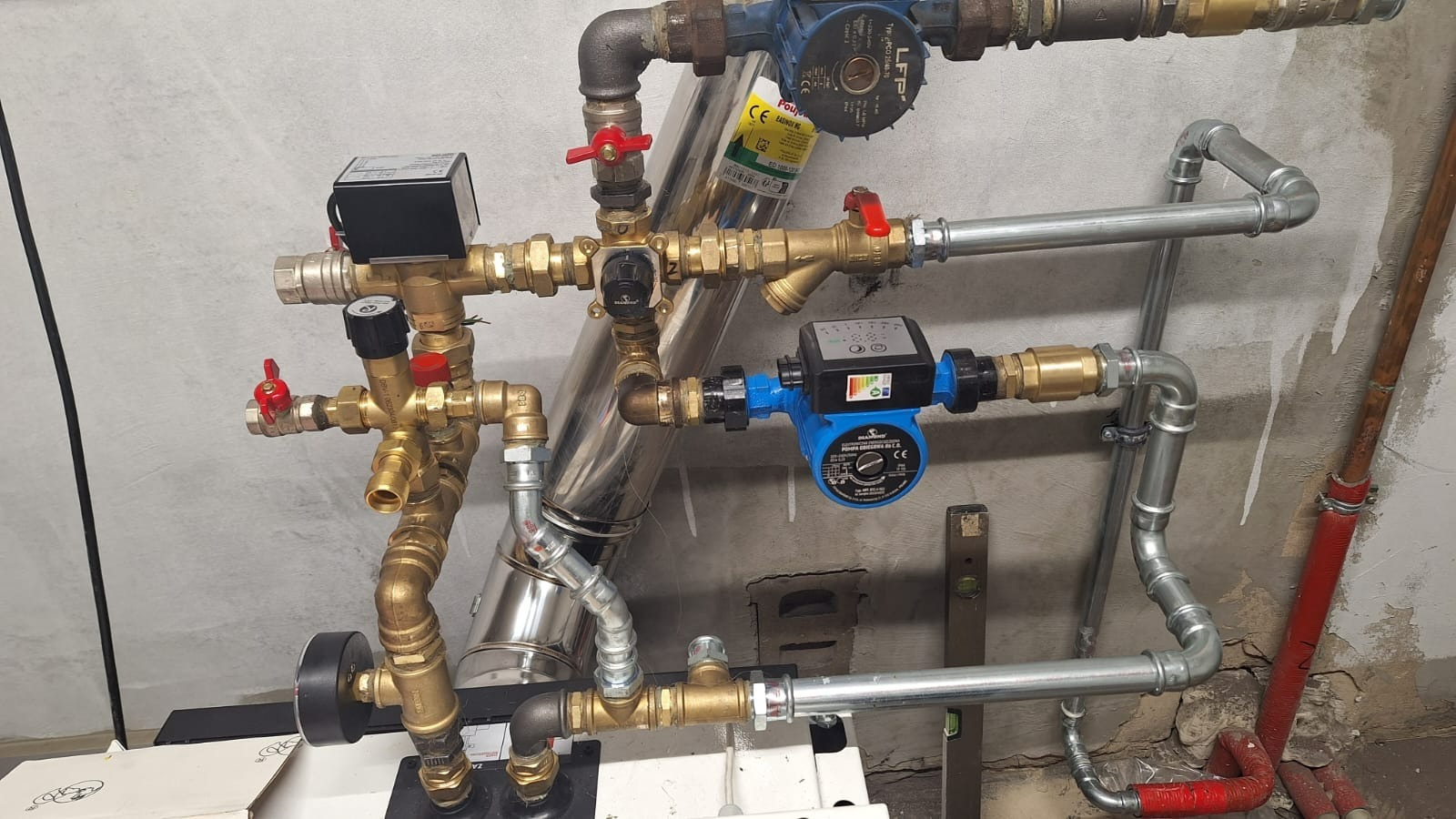 Złożony układ rur i zaworów z pompami obiegowymi na tle surowej ściany z widocznymi śladami napraw