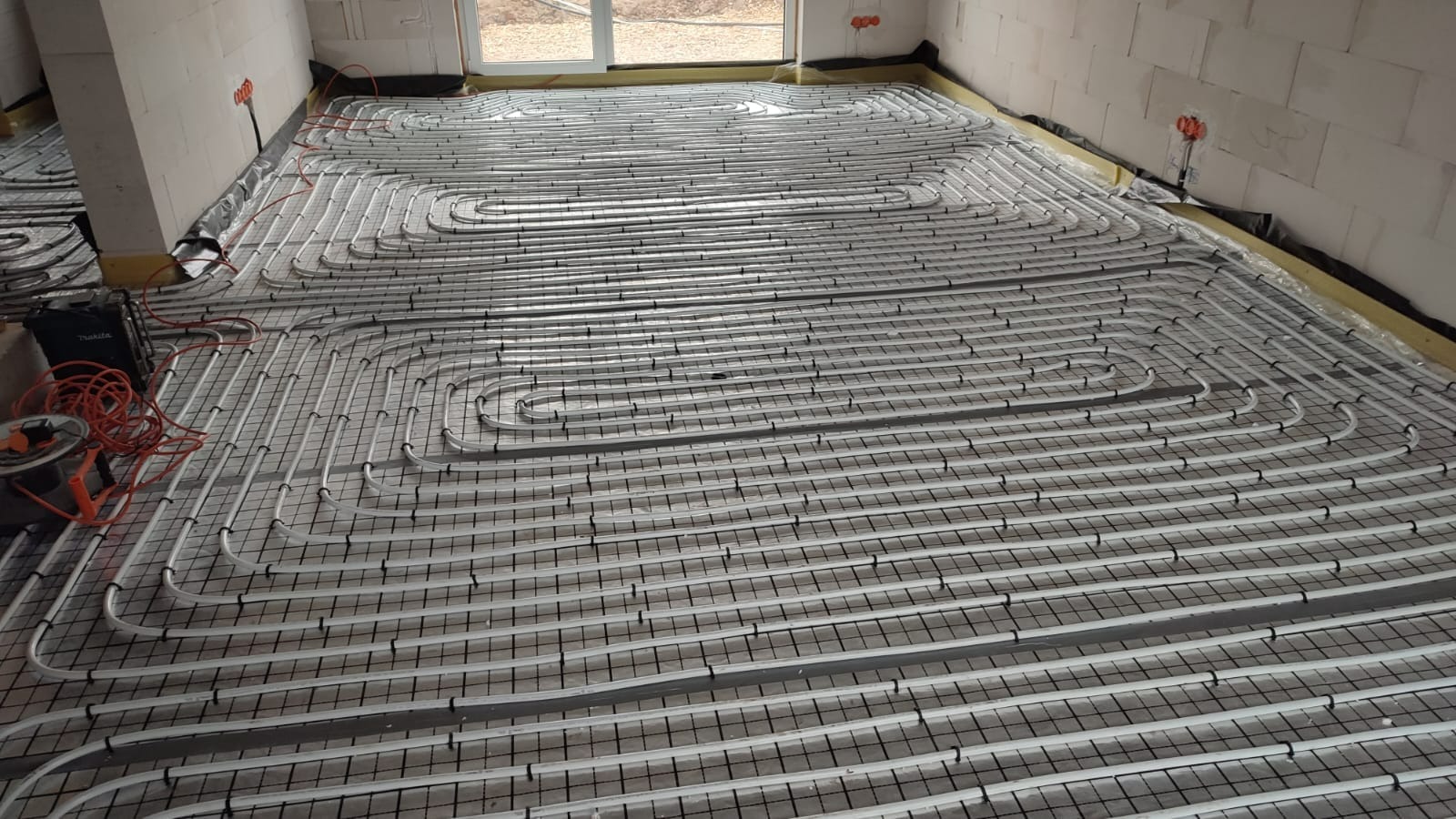 Białe rury ogrzewania podłogowego zamocowane na czarnej siatce w surowym stanie deweloperskim, widoczne narzędzia montażowe w rogu pomieszczenia.
