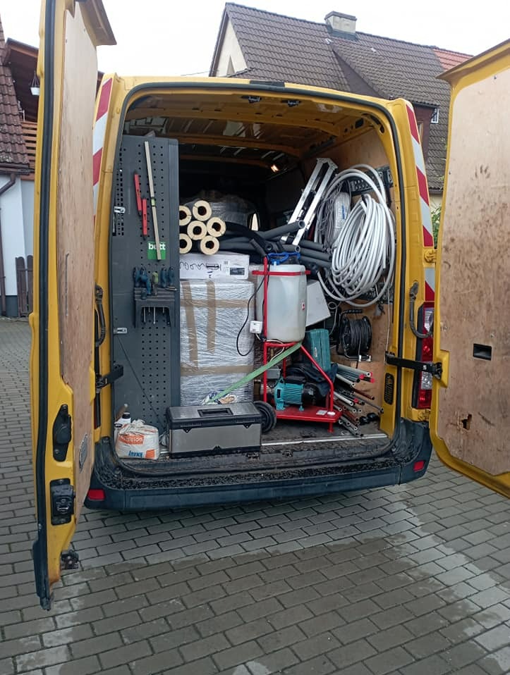 Wnętrze żółtego samochodu dostawczego z otwartymi drzwiami, wypełnione narzędziami i materiałami instalacyjnymi, w tle budynki mieszkalne.