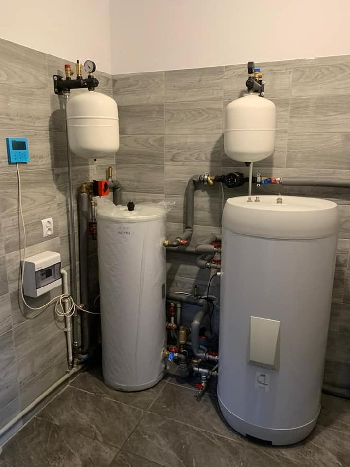 Dwa białe zbiorniki akumulacyjne z osprzętem hydraulicznym w pomieszczeniu wyłożonym płytkami imitującymi drewno, widoczna skrzynka elektryczna i regulator temperatury na ścianie.