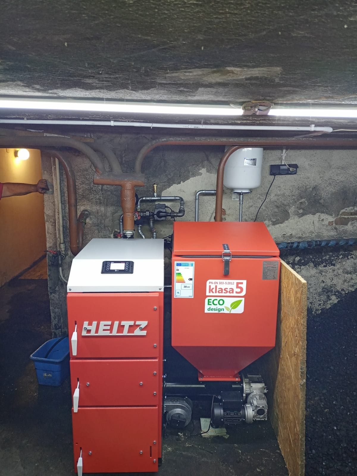 Nowoczesny, czerwony piec na paliwo stałe marki HEITZ z podajnikiem, zainstalowany w piwnicy z widocznymi rurami i instalacją grzewczą przy ścianie z surowego betonu. Piec oznaczony etykietą klasy...