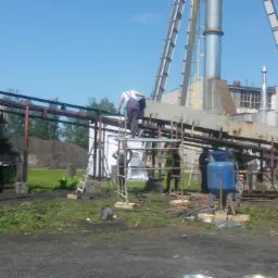 Prace przygotowawcze do piaskowania konstrukcji przemysłowej w Leżajsku: pracownik na rusztowaniu, agregat piaskarski i zbiornik, widoczne ślady korozji na elementach.