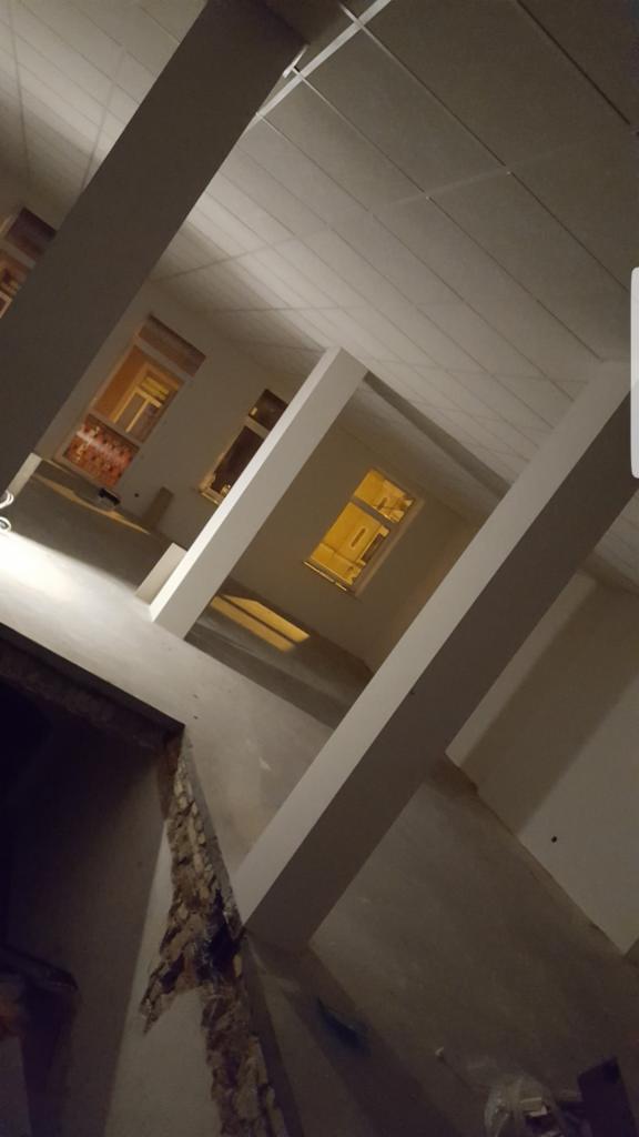 Wnętrze w trakcie remontu z widocznymi elementami konstrukcyjnymi, oknami z widokiem na oświetlone budynki i fragmentem sufitu podwieszanego.