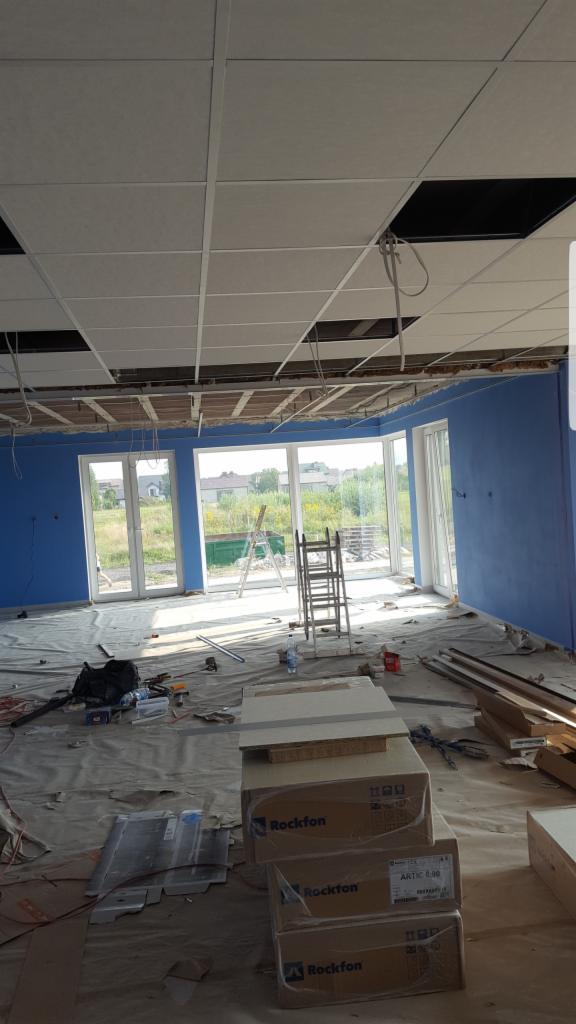 Montaż sufitu podwieszanego z widocznymi elementami konstrukcyjnymi i otworami na instalacje, niebieskie ściany, okna z widokiem na zewnątrz, podłoga zabezpieczona folią, kartony z materiałami...