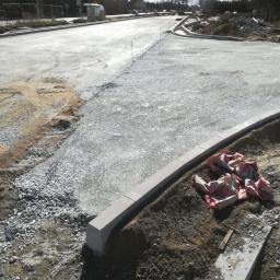 BET brukarstwo maszynowe - przemysłowe - Budowa nowej drogi: widok na przygotowany podkład z kruszywa, ustawione krawężniki betonowe, w tle zabudowa miejska i słup oświetleniowy.