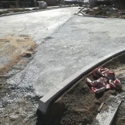 Budowa nowej drogi: widok na przygotowany podkład z kruszywa, ustawione krawężniki betonowe, w tle zabudowa miejska i słup oświetleniowy.