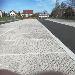 BET brukarstwo maszynowe - przemysłowe - Rozległy parking z ażurowej kostki betonowej wypełnionej drobnym kruszywem, oddzielony pasem asfaltu, w tle budynki mieszkalne pod błękitnym niebem.