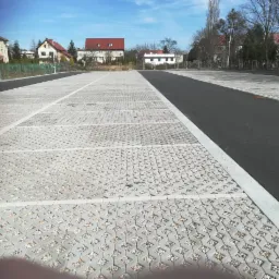 Rozległy parking z ażurowej kostki betonowej wypełnionej drobnym kruszywem, oddzielony pasem asfaltu, w tle budynki mieszkalne pod błękitnym niebem.