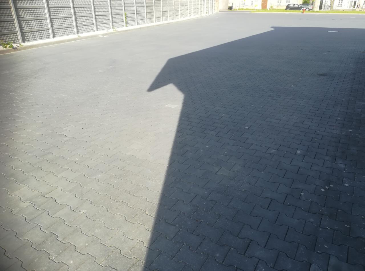 Ułożona, szara kostka brukowa na dużym placu, z widocznym cieniem budynku i fragmentem ogrodzenia akustycznego w tle.