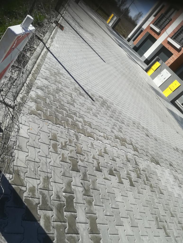 Nowo ułożona, szara kostka brukowa w kształcie litery H, widoczny fragment budynku z żółtymi elementami i biało-czerwony słupek zabezpieczający.