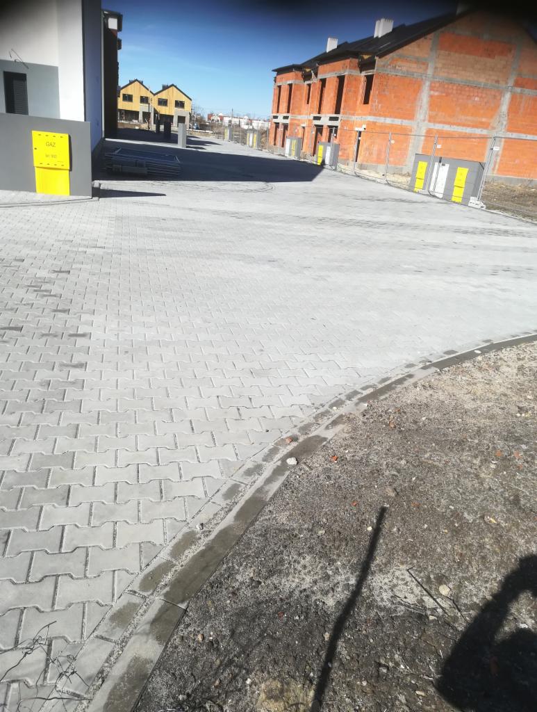 Ułożona szara kostka brukowa na podjeździe przed nowymi budynkami mieszkalnymi, widoczna skrzynka gazowa przy budynku.