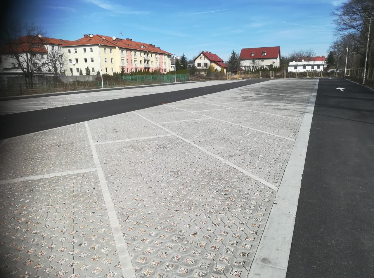Nowy parking z kostki ażurowej w jasnoszarym odcieniu, oddzielony pasem asfaltu, z budynkami mieszkalnymi w tle, oświetlony jasnym, dziennym światłem.