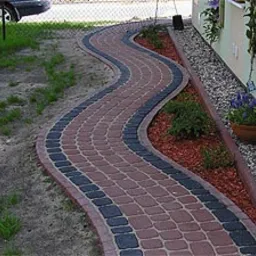 Kręta ścieżka ułożona z czerwono-czarnej kostki prowadząca wzdłuż budynku z kwiatami w donicach, otoczona trawnikiem i rabatą z czerwoną korą.