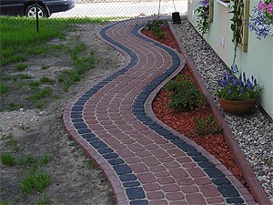 Kręta ścieżka ułożona z czerwono-czarnej kostki prowadząca wzdłuż budynku z kwiatami w donicach, otoczona trawnikiem i rabatą z czerwoną korą.